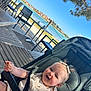 baby, bow_hair, buildings, cheeks, grass, hand, infant, lake, outdoor, patio_chairs, pine_tree, seat, sky, smiling, stroller, sunny, table, townscape, waterfront, wooden_deck