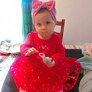 Shayna a rejoint le concours — aidez-le/la à gagner de superbes lots ! child, toddler, baby, pink_headband, bow, red_dress, sitting, chair, toy, indoor, curtain, window, portrait_photo, table, shoes, socks, blanket, hand, face, cute