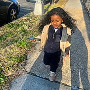 Luis is registered to the contest to win money with this photo: child, curly_hair, sidewalk, smartphone, jacket, urban, street, sunlight, shadow, car, tree, grass, walking, person, casual_clothing, outdoor, daytime, cool, confident, portrait