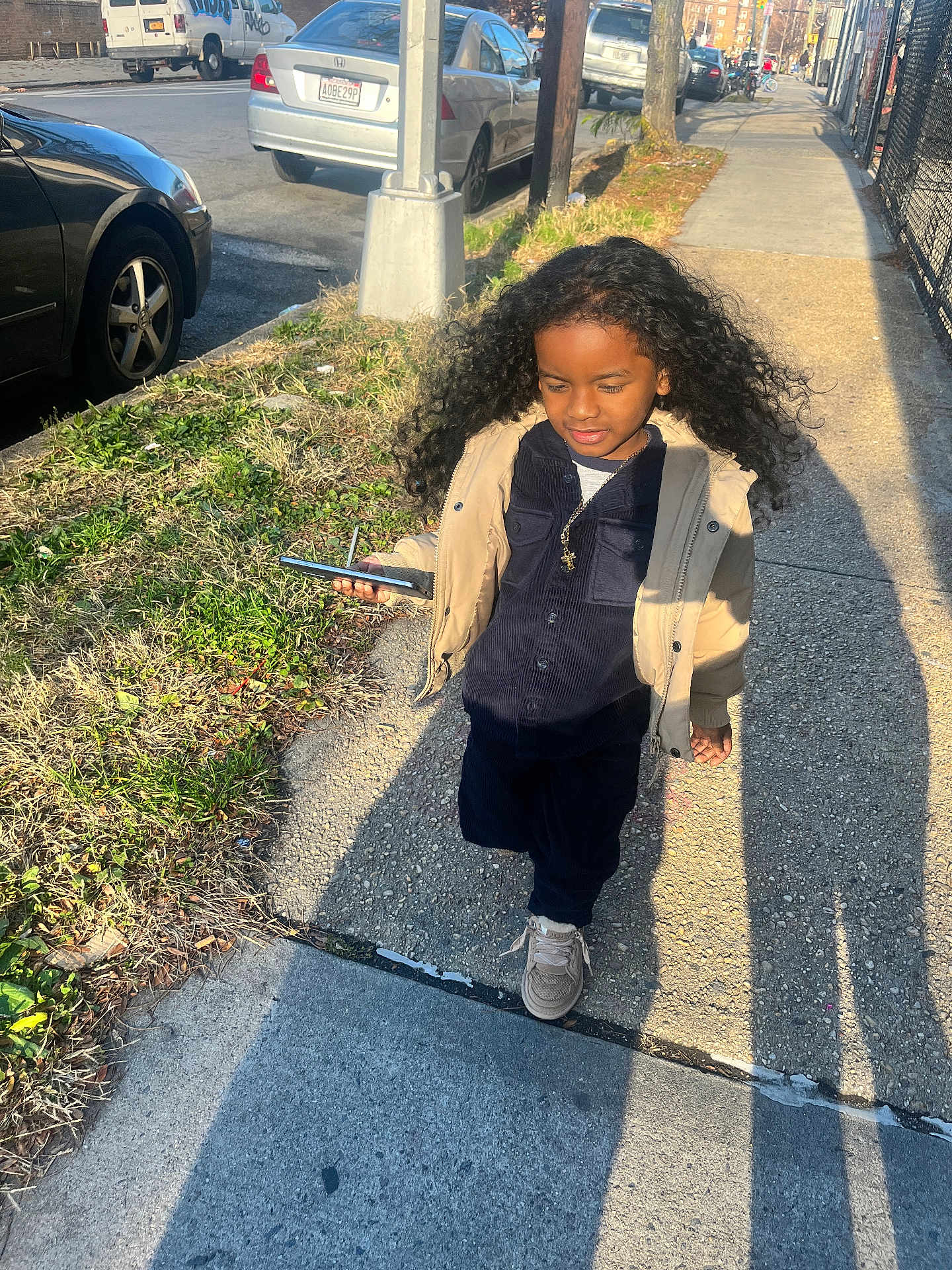 Luis is registered to the contest to win money with this photo: child, curly_hair, sidewalk, smartphone, jacket, urban, street, sunlight, shadow, car, tree, grass, walking, person, casual_clothing, outdoor, daytime, cool, confident, portrait