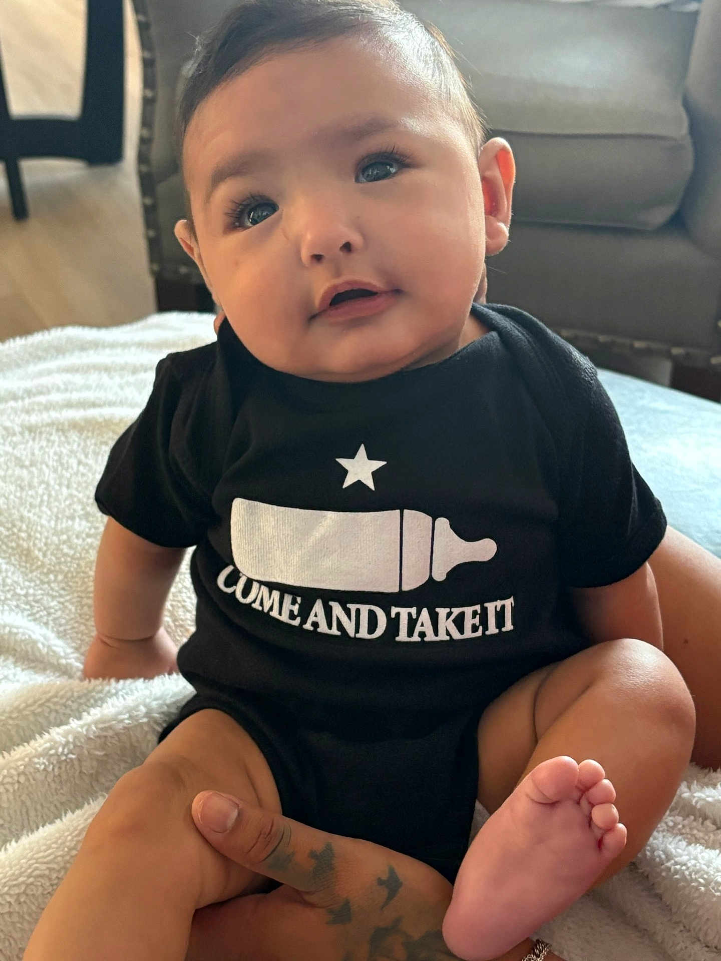 Aurelius is registered to the contest to win money with this photo: baby, infant, child, onesie, black_clothing, bottle_graphic, text_on_clothing, tattoo, hand, foot, blanket, indoor, furniture, couch, floor, face, portrait, cute, sitting, soft_light