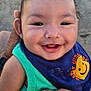 baby, smiling, child, green_shirt, bib, happy, hand, tattoo, outdoor, sand, face, portrait, cute, infant, person, clothing, skin, closeup, expression, joy