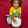 child, toddler, curly_hair, portrait, sitting, tambourine, musical_instrument, drumming, yellow_shirt, animal_print, gray_pants, sneakers, red_carpet, indoors, looking_at_camera, serious_expression, playful, music, portrait_photography, colorful