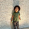 casual_clothing, child, concrete, curly_hair, floor, full_body, green_shirt, jeans, natural_light, outdoor, photography, portrait, shadow, smiling, sneakers, standing, textured_wall, toddler, toy, wall
