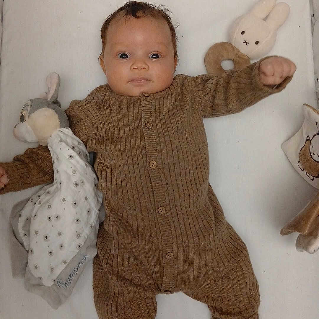 Isayah participe au concours pour gagner de l'argent avec cette photo : baby, blanket, brown_onesie, child, comfort, cozy, cute, expression, face, hand, indoors, infant, knitted_clothing, lying_down, person, plush_toy, sock, soft_toy, stuffed_animal, white_background