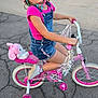 child, bicycle, helmet, pink, denim_overalls, training_wheels, plush_toy, outdoor, sidewalk, smiling, happy, road, toy, casual_clothing, playful, sunlight, fun, transportation, helmet_unicorn, young