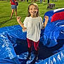 child, smiling, inflatable, playground, night, outdoor, event, people, crowd, food_truck, grass, blue, white_shirt, red_pants, fun, happy, festival, family, recreation, leisure