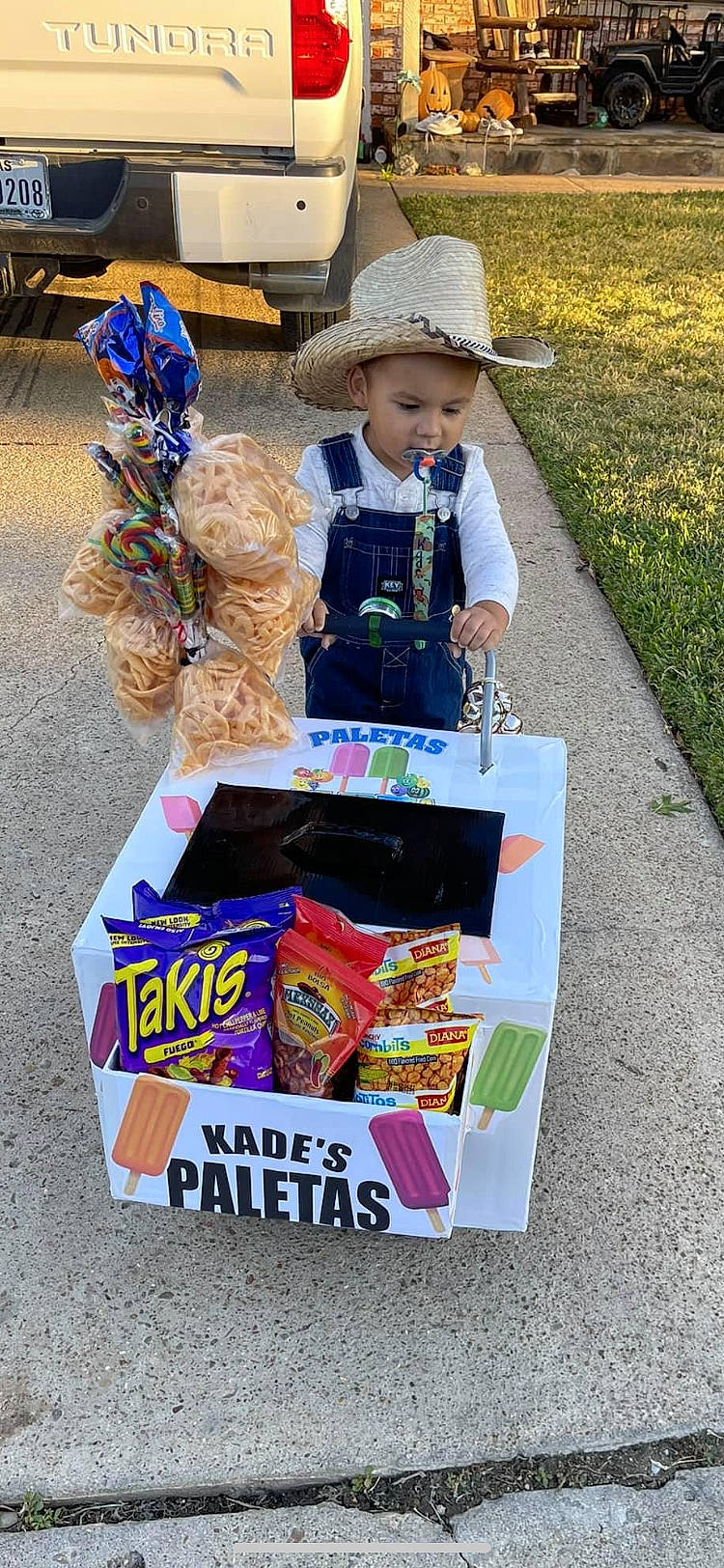 Kade is registered to the contest to win money with this photo: advertising, asphalt, baby, box, carton, child, event, fun, grass, hat, headwear, packaging_and_labeling, person, plant, public_event, recreation, soil, summer, sun_hat, t_shirt