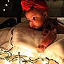 baby, child, red_headband, flower_headband, string_lights, blanket, soft_lighting, indoors, curious, infant, closeup, warm_tone, candid, portrait, cozy, holiday_lights, face, hand, expression, decor