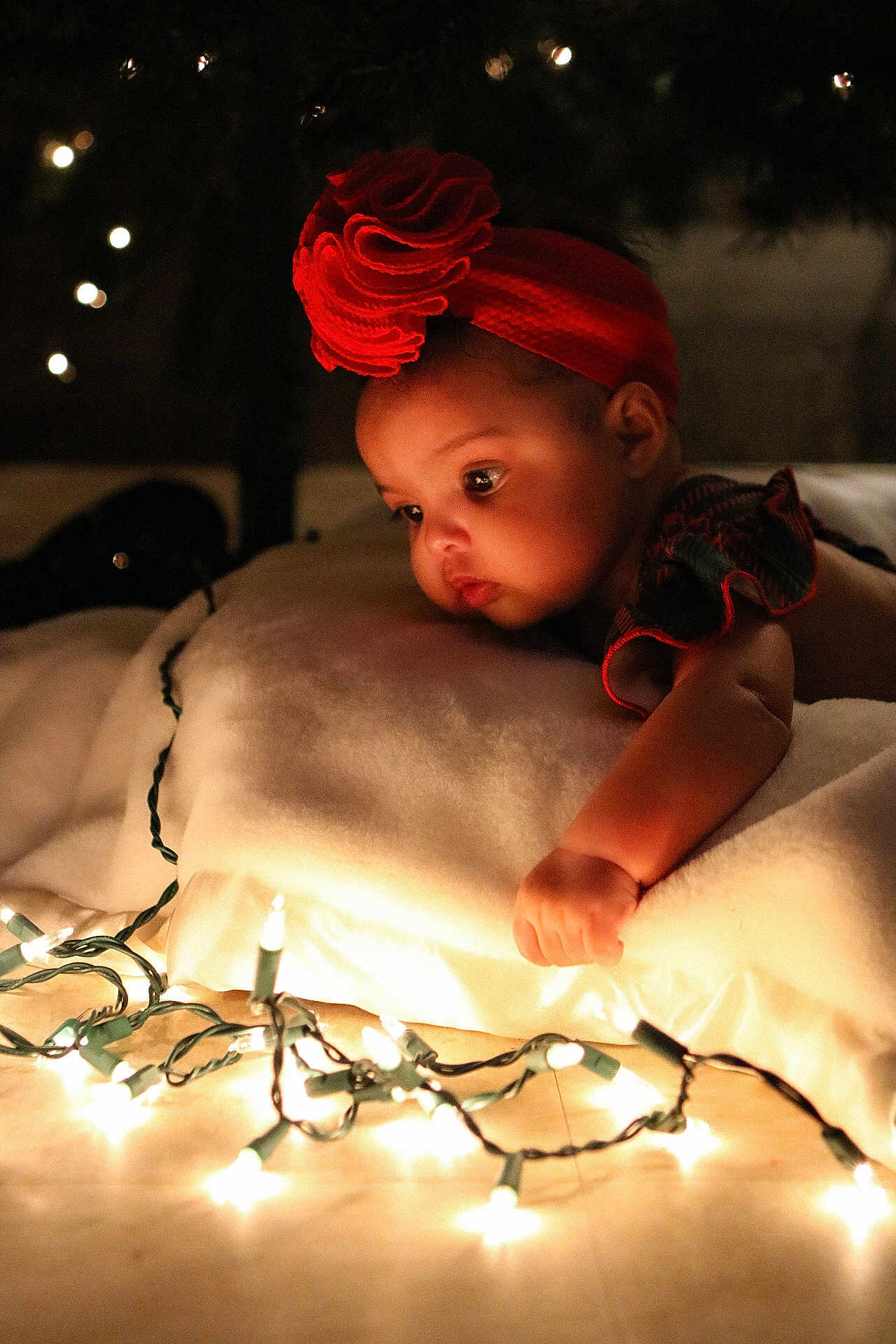 Ava is registered to the contest to win money with this photo: baby, child, red_headband, flower_headband, string_lights, blanket, soft_lighting, indoors, curious, infant, closeup, warm_tone, candid, portrait, cozy, holiday_lights, face, hand, expression, decor