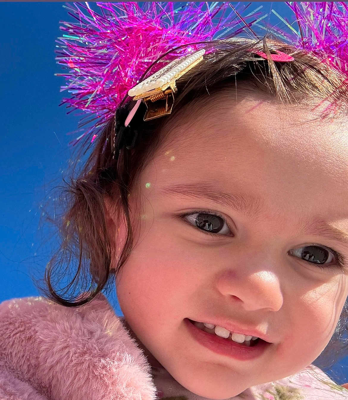 November is registered to the contest to win money with this photo: child, toddler, portrait, close_up, smile, teeth, eye, hair, headband, hair_clip, tinsel, pink, fuzzy_coat, jacket, outdoor, blue_sky, sunlight, reflection, cute, happy