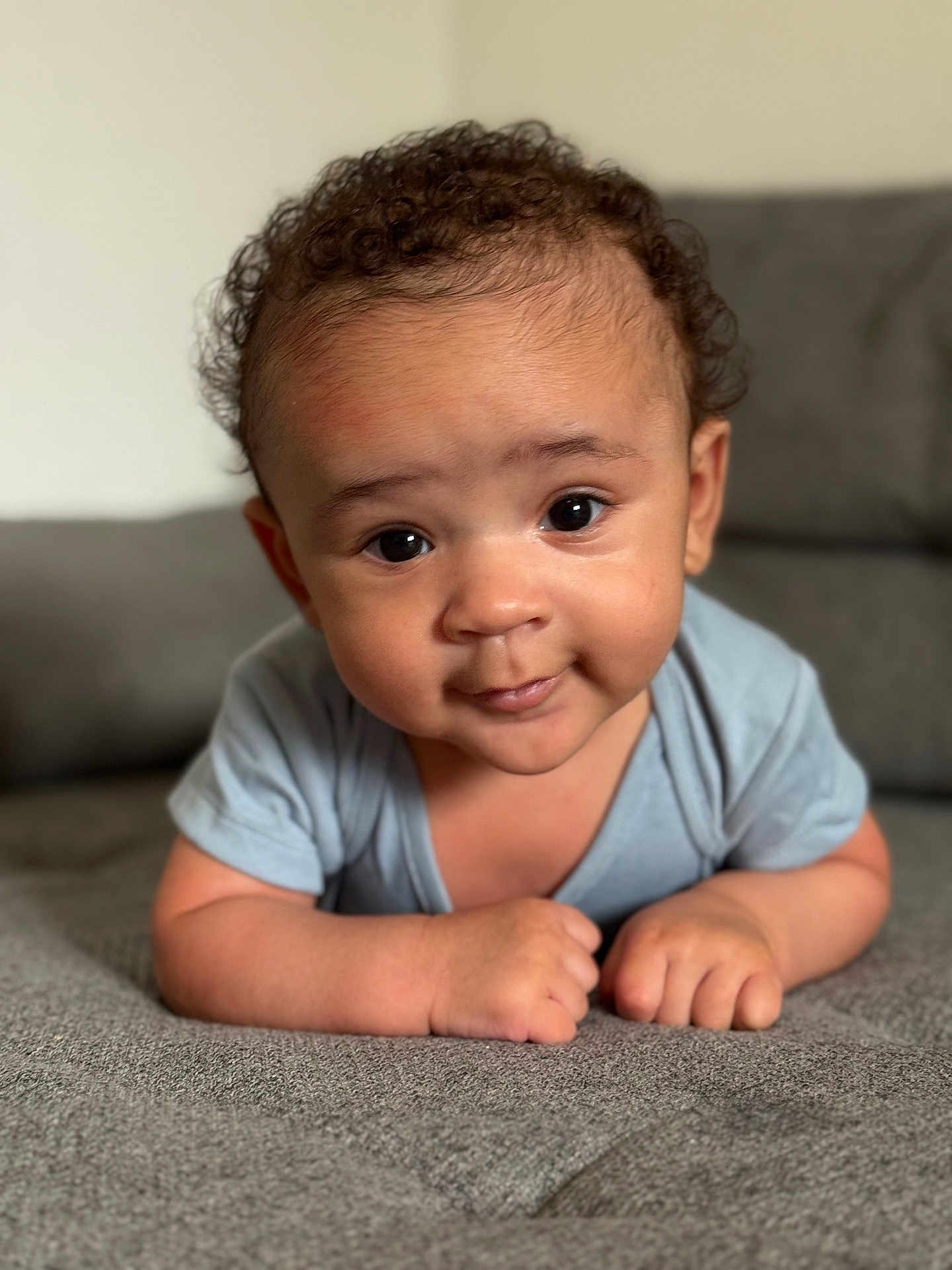 Zyon is registered to the contest to win money with this photo: baby, infant, child, face, eyes, smile, curly_hair, couch, gray, skin, hands, short_sleeve, clothing, indoors, portrait, cute, young, person, toddler, expression