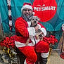 man, dog, santa_claus_costume, glasses, beard, holiday_decorations, poinsettia, reindeer_sleigh, indoor, smiling, festive, pet_smart_sign, red_clothing, black_boots, leash, seated, christmas_theme, background, portrait, celebration
