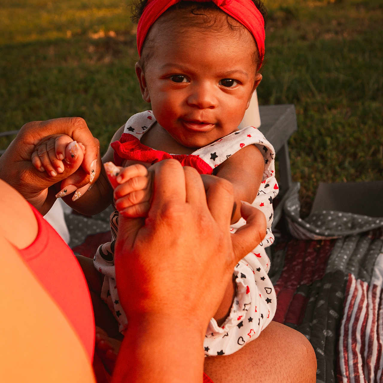 Sianni is registered to the contest to win money with this photo: adult_hand, baby, blanket, child, cute, family, grass, holding_hands, infant, nature, outdoor, patterned_clothing, person, picnic, playful, red_headband, smiling, sunlight, sunset, toddler