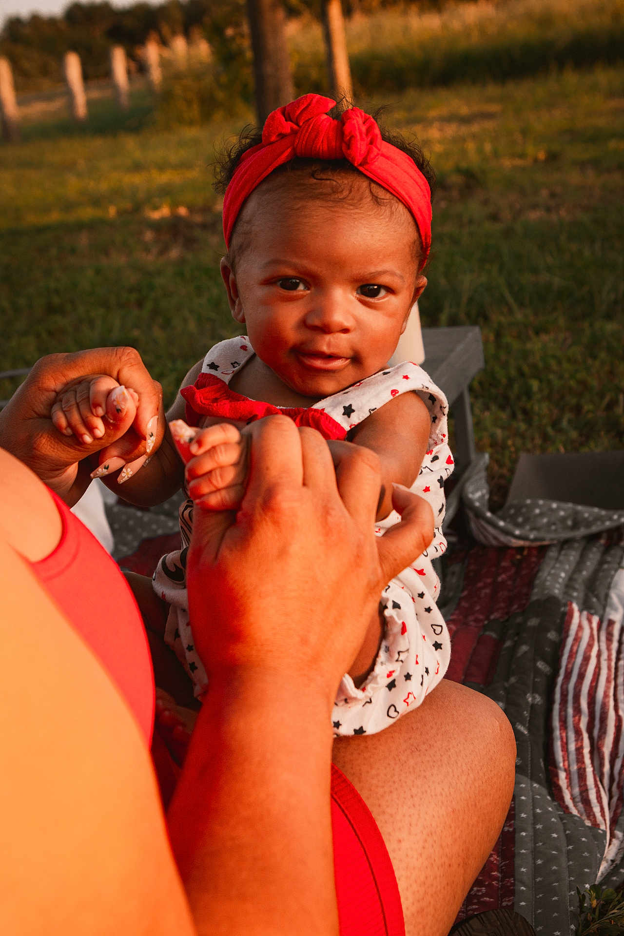 Sianni is registered to the contest to win money with this photo: baby, child, red_headband, holding_hands, outdoor, picnic, blanket, grass, sunset, playful, person, cute, infant, smiling, nature, sunlight, family, toddler, patterned_clothing, adult_hand
