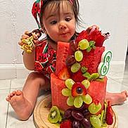 Duleivis Isabella is registered to the contest to win money with this photo: toddler, child, fruit_cake, watermelon, grapes, strawberries, kiwi, toy_doll, headband, red_clothing, indoor, floor, curious_expression, barefoot, cake, mint_leaves, wooden_plate, playful, colorful, cute