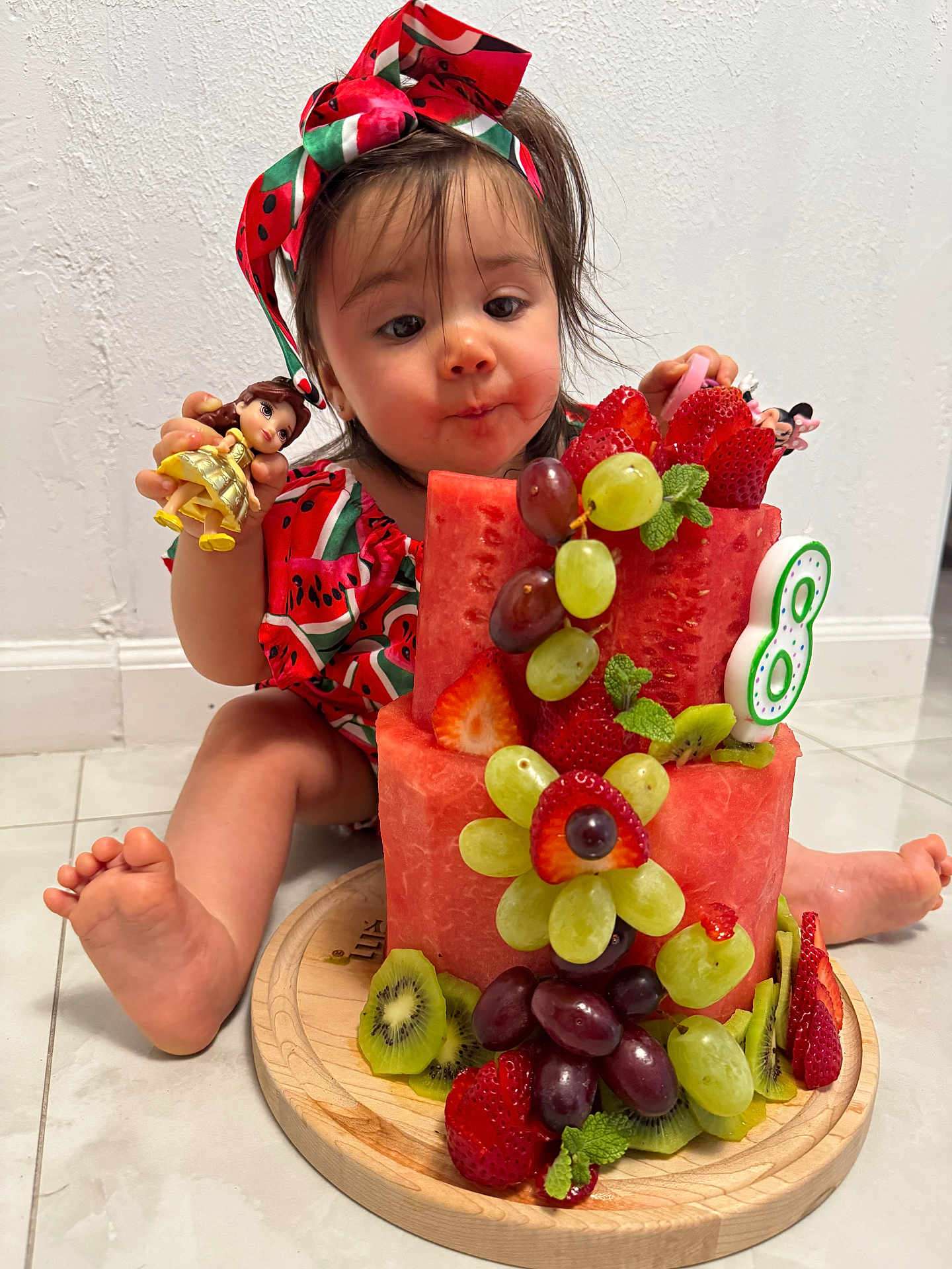 Duleivis Isabella is registered to the contest to win money with this photo: toddler, child, fruit_cake, watermelon, grapes, strawberries, kiwi, toy_doll, headband, red_clothing, indoor, floor, curious_expression, barefoot, cake, mint_leaves, wooden_plate, playful, colorful, cute