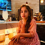 Selena is registered to the contest to win money with this photo: child, girl, dress, pink_dress, sitting, chair, indoor, modern_interior, lighting, glass, lemonade, drink, television, underwater_scene, hair, long_hair, face, expression, warm_light, bar_counter