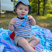 Beau joined the competition — help win amazing prizes! baby, child, blue_hat, striped_shirt, teething_toy, blanket, outdoor, nature, grass, trees, toy, sitting, curious, infant, daylight, person, closeup, playtime, casual_clothing, relaxation