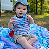 baby, child, blue_hat, striped_shirt, teething_toy, blanket, outdoor, nature, grass, trees, toy, sitting, curious, infant, daylight, person, closeup, playtime, casual_clothing, relaxation