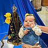 Darvy Domingos Veiga is registered to the contest to win money with this photo: adult, baby, beige_pants, blue_clothing, blue_drapery, ceremonial, child, crown, flowers, hand, indoor, infant, looking_at_camera, portrait, prayer_beads, religious_statue, roses, safe, sitting, vest