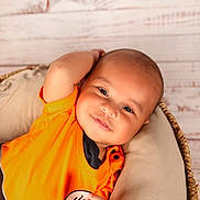William is registered to the contest to win money with this photo: baby, infant, orange_clothing, smile, face, head, arm, cushion, basket, wooden_background, relaxing, portrait, child, cute, skin, indoors, comfortable, happy, person, casual