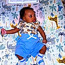 animal_print, baby, blanket, blue_shorts, book, child, colorful, curious, cute, elephant, giraffe, indoor, infant, lying_down, open_book, patterned_socks, plants, playful, soft_lighting, young_child