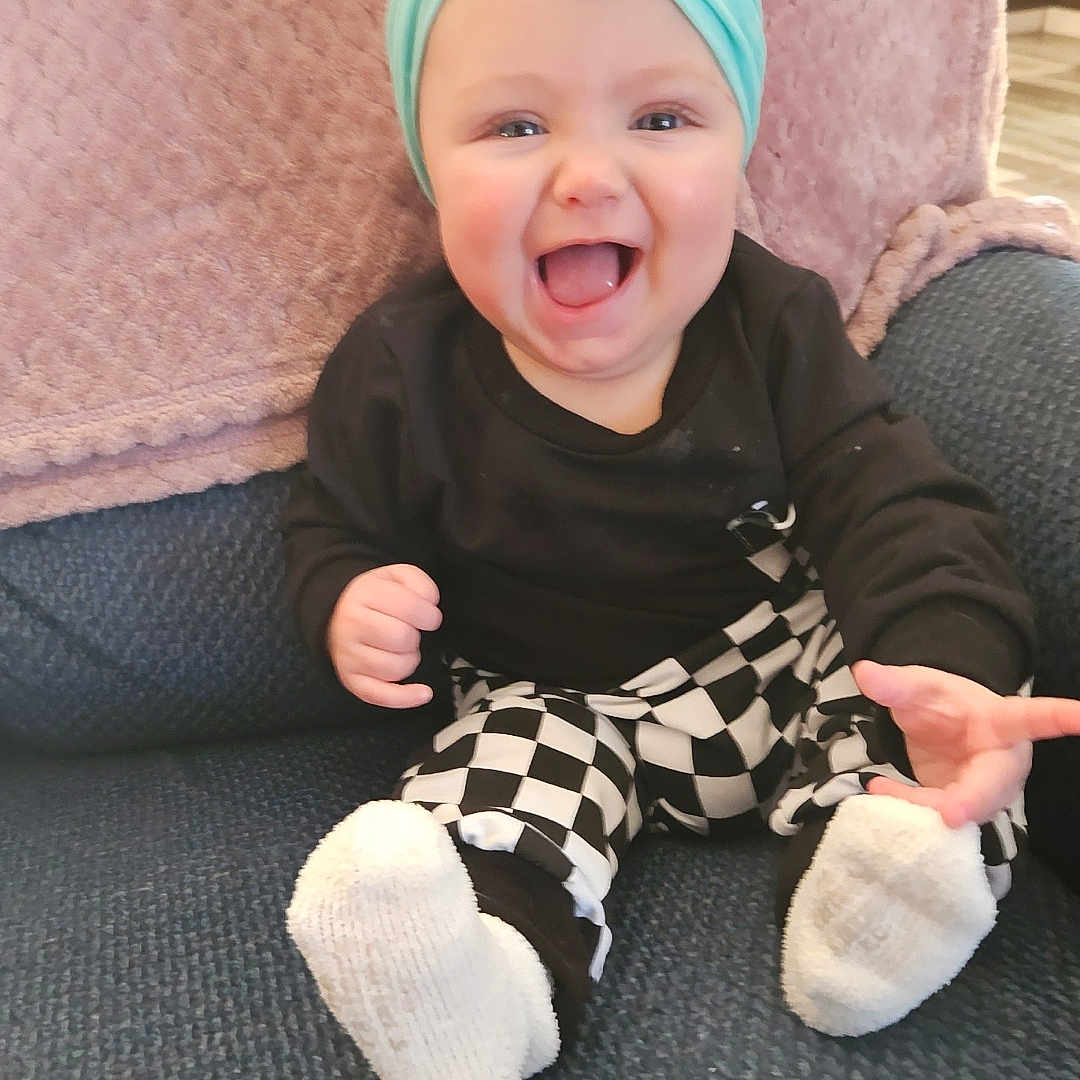 Emma is registered to the contest to win money with this photo: baby, cap, clothing, couch, cushion, face, footwear, furniture, happy, hat, head, homedecor, hosiery, laughing, pants, person, photography, portrait, sitting, sock
