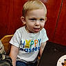 boy, casual_clothing, chair, child, curious_expression, easter, family, food, happy_easter_shirt, indoor, meal, messy_face, person, plate, portrait, short_blond_hair, sitting, table, toddler, wooden_background