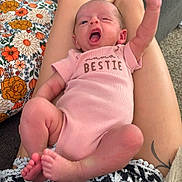 Ariella joined the competition — help win amazing prizes! baby, infant, pink_onesie, raised_fist, legs, tattoo, sofa, blanket, floral_pillow, carpet, person, skin, feet, living_room, cozy, expression, cute, newborn, relaxed, home