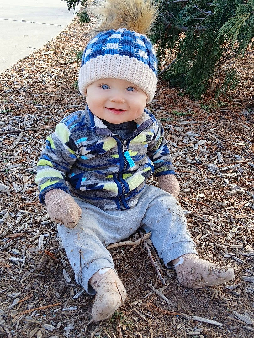 Jade is registered to the contest to win money with this photo: blue_eyes, casual_clothing, child, daylight, dirt, evergreen, face, happy, jacket, knit_hat, nature, outdoor, pants, person, playful, pom_pom, sitting, smiling, toddler, wood_chips