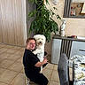 woman, person, dog, white_dog, pet, hugging, kneeling, smiling, portrait, indoors, living_room, home, houseplant, mirror, reflection, furniture, sideboard, chair, tiled_floor, wall_decor