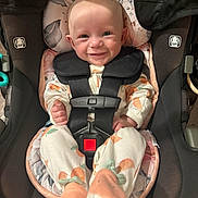 June is registered to the contest to win money with this photo: apparel, baby, car_seat, child, cute, face, footwear, happy, head, indoor, infant, onesie, orange, patterned_clothing, person, plush_slippers, safety_harness, seat, smile, straps