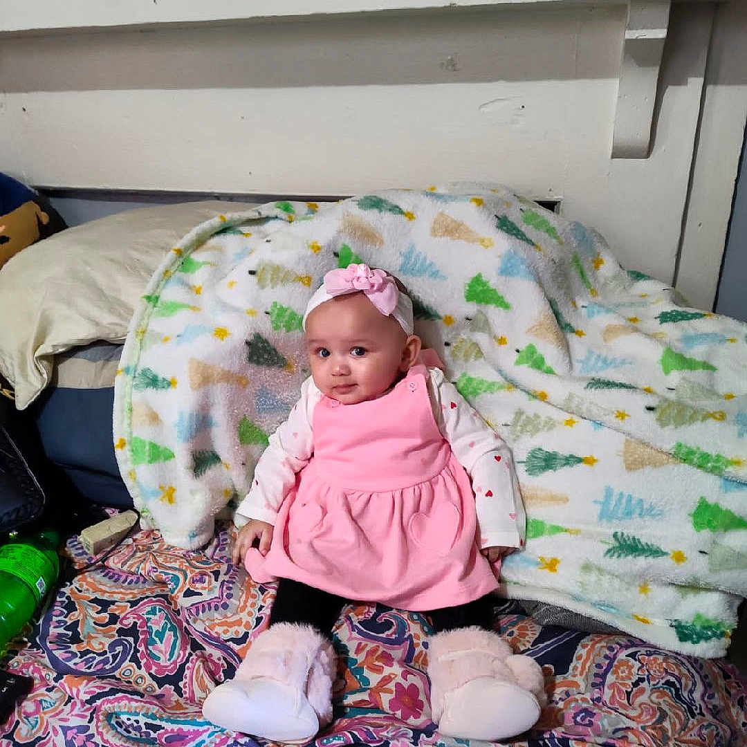 Journey is registered to the contest to win money with this photo: baby, blanket, child, colorful, cozy, curious, cute, face, furniture, headband, home, household_items, indoor, patterned_bedspread, person, pink_dress, portrait, shelf, sitting, slippers