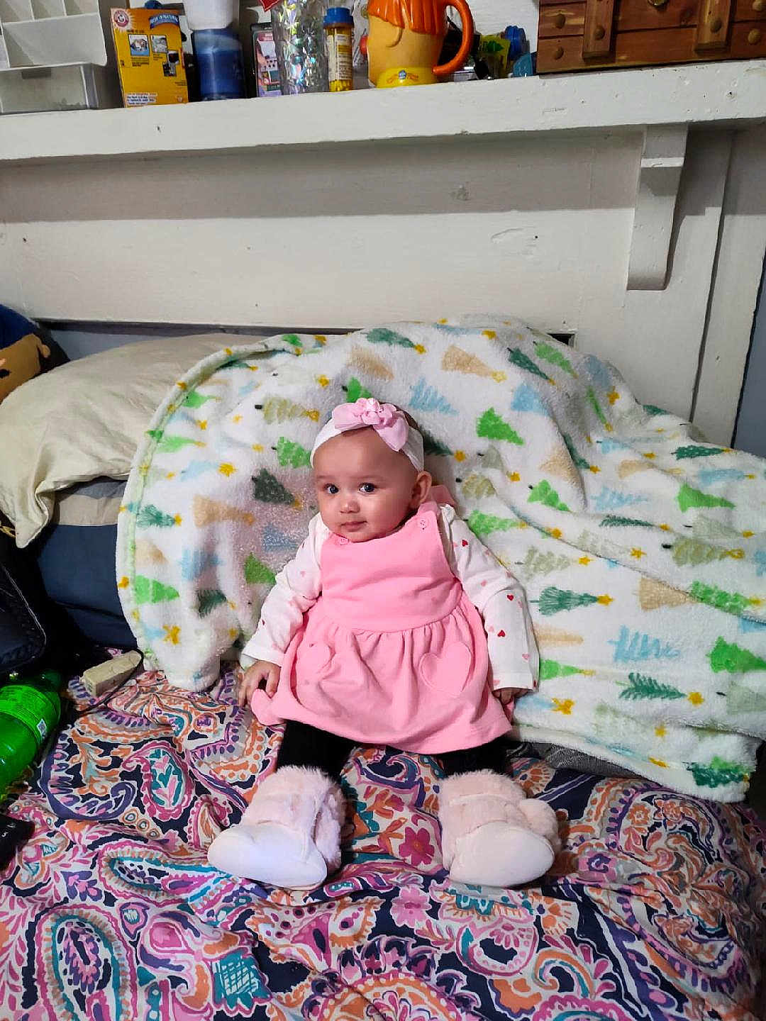 Journey is registered to the contest to win money with this photo: baby, pink_dress, headband, slippers, blanket, patterned_bedspread, indoor, child, cute, sitting, portrait, cozy, furniture, home, shelf, household_items, curious, face, person, colorful