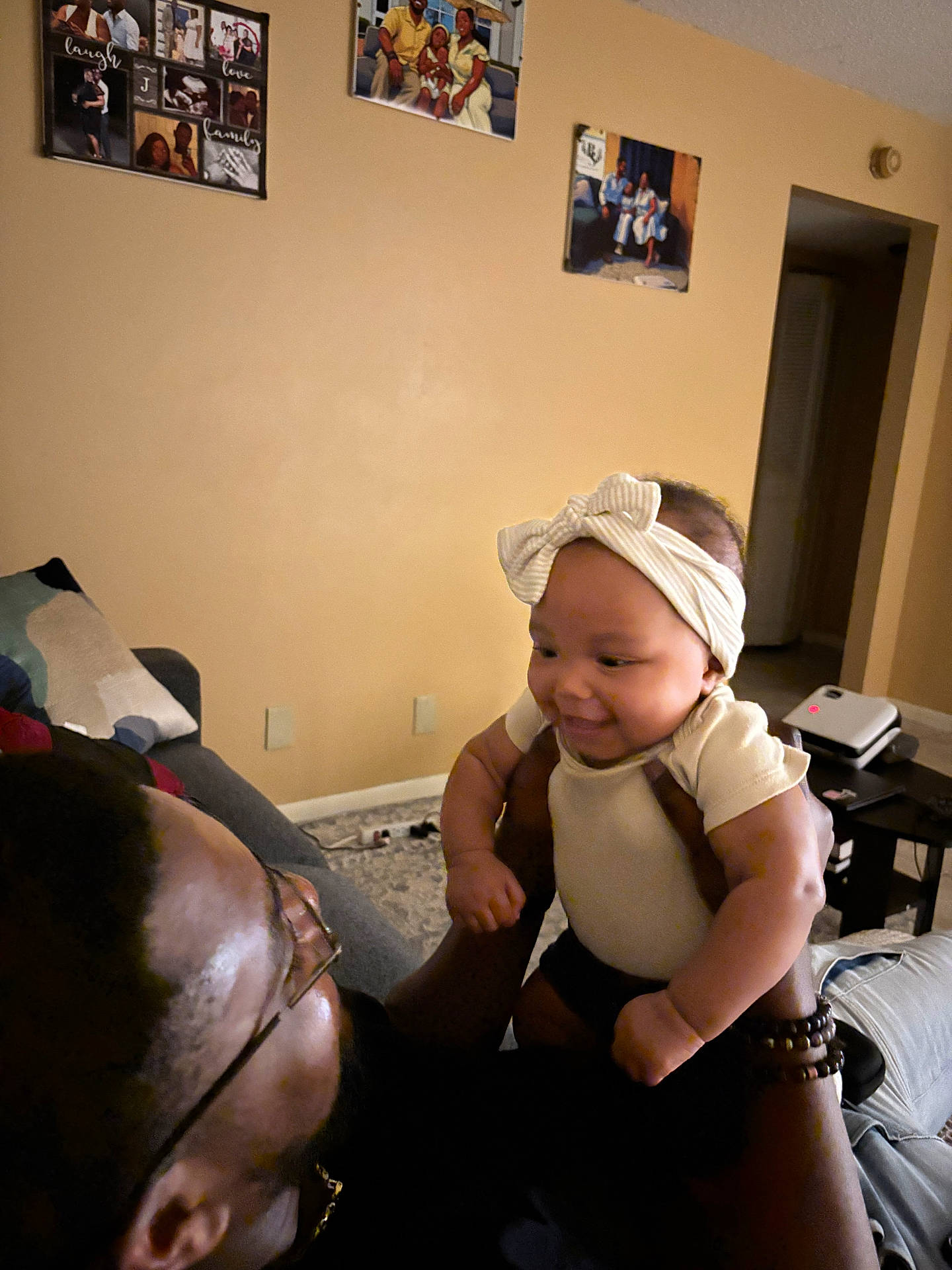 adult, baby, bracelet, carpet, clothing, couch, device, family_photos, glasses, happy, headband, holding, home, indoor, interaction, living_room, person, smile, table, wall_art