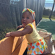 Tiana is registered to the contest to win money with this photo: bag, bench, child, colorful, curious, daytime, fence, headband, outdoor, patterned_clothing, person, playground, shadow, skirt, standing, sunlight, tennis_court, toddler, yellow_top, young_child