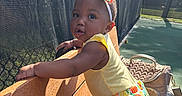 Tiana is registered to the contest to win money with this photo: bag, bench, child, colorful, curious, daytime, fence, headband, outdoor, patterned_clothing, person, playground, shadow, skirt, standing, sunlight, tennis_court, toddler, yellow_top, young_child