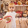 blonde_hair, blue_eyes, candy_cane, child, christmas_theme, cute, festive, fluffy_decoration, gift_box, holiday_decor, holiday_spirit, indoor, portrait, red_ribbon, stuffed_animal, teddy_bear, toddler, white_sweater, winter_clothing, wooden_floor