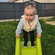 Laÿna participe au concours pour gagner de l'argent avec cette photo : toddler, child, baby, slide, green_slide, outdoor, backyard, grass, pigtails, vest, sneakers, smile, face, hands, standing, playground, fence, house, portrait, young_child
