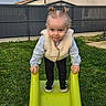 baby, backyard, child, face, fence, grass, green_slide, hands, house, outdoor, pigtails, playground, portrait, slide, smile, sneakers, standing, toddler, vest, young_child