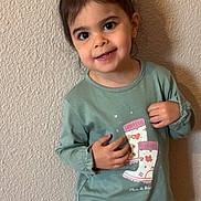 Séréna participe au concours pour gagner de l'argent avec cette photo : child, toddler, girl, smile, portrait, standing, green_shirt, boot_graphic, wall, textile, ponytail, hair_tie, big_eyes, earring, teeth, hands, indoor, cute, casual_clothing, shadow