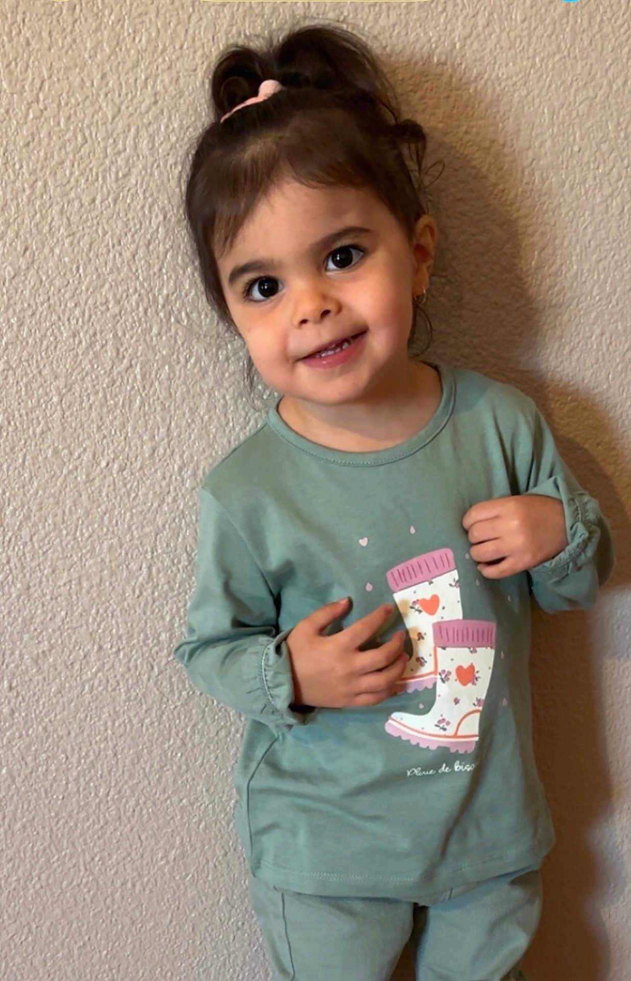 Séréna participe au concours pour gagner de l'argent avec cette photo : child, toddler, girl, smile, portrait, standing, green_shirt, boot_graphic, wall, textile, ponytail, hair_tie, big_eyes, earring, teeth, hands, indoor, cute, casual_clothing, shadow