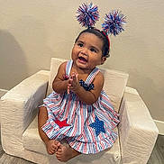 Aamiyah is registered to the contest to win money with this photo: baby, child, clapping, dress, headband, pom_poms, armchair, indoor, floor, wall, happy, smiling, footwearless, star_pattern, red, white, blue, person, portrait, cute