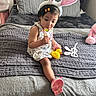 toddler, child, bunny_ears, lollipop, yellow_rubber_duck, bed, blanket, pillow, toy, bracelet, hair_clip, indoor, cute, pink_shoe, dress, person, soft_toys, playful, cozy, smiling