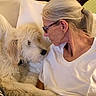 affection, bed, blanket, blonde_hair, closeup, cozy, cuddling, dog, face, fluffy_fur, glasses, indoors, person, pet, pillows, ponytail, portrait, warm_lighting, white_shirt, woman