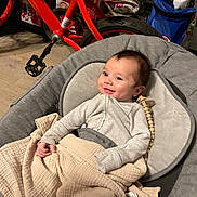 Cameron is registered to the contest to win money with this photo: baby, smiling, blanket, gray_cushion, red_bike, indoor, floor, toy, child, person, clothing, seat, wooden_floor, home, smile, infant, comfort, happy, casual, relaxing