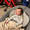 baby, smiling, blanket, gray_cushion, red_bike, indoor, floor, toy, child, person, clothing, seat, wooden_floor, home, smile, infant, comfort, happy, casual, relaxing