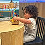 toddler, child, curly_hair, playing, toy, wooden_table, bead_maze, indoor, chair, black_chair, casual_clothing, white_shirt, gray_overall, focus, side_view, wood_texture, flooring, aquarium, background_blur, person