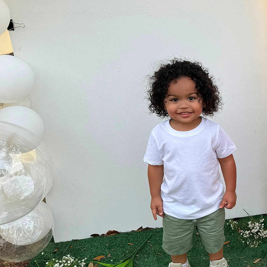 Azhainei joined the competition — help win amazing prizes! background, balloons, casual_clothing, celebration, child, curly_hair, cute, flowers, green_grass, green_shorts, happy, nature, outdoor, person, portrait, smiling, sneakers, standing, toddler, white_tshirt